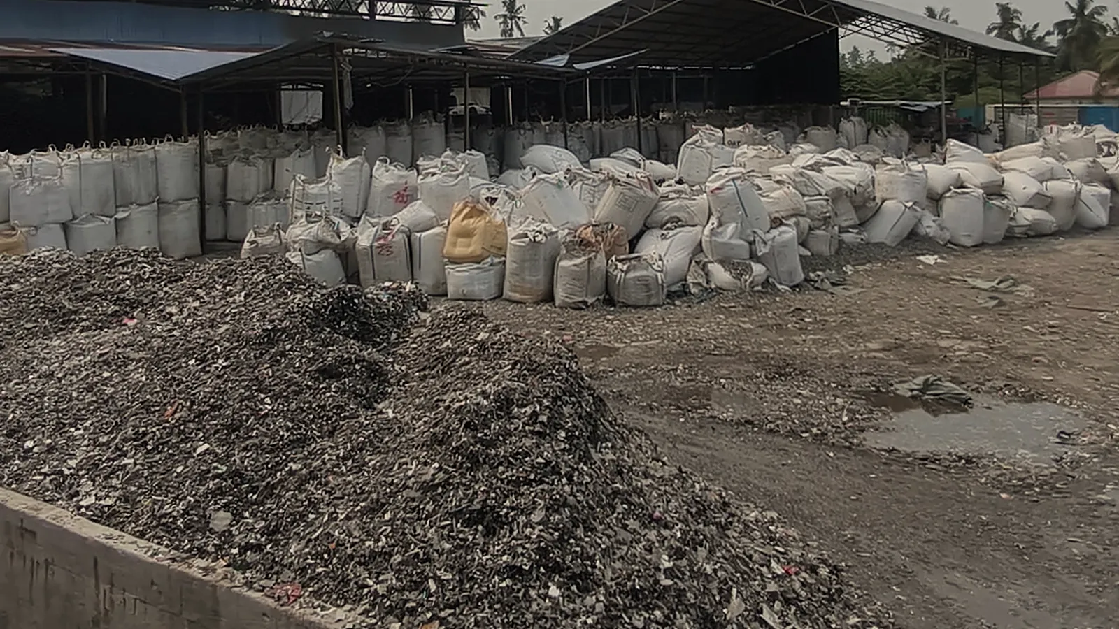 Crushed plastic scrap heaped in an open yard beside a low concrete wall, with rows of bulk bags beyond, in Dayatas Profab's production footage