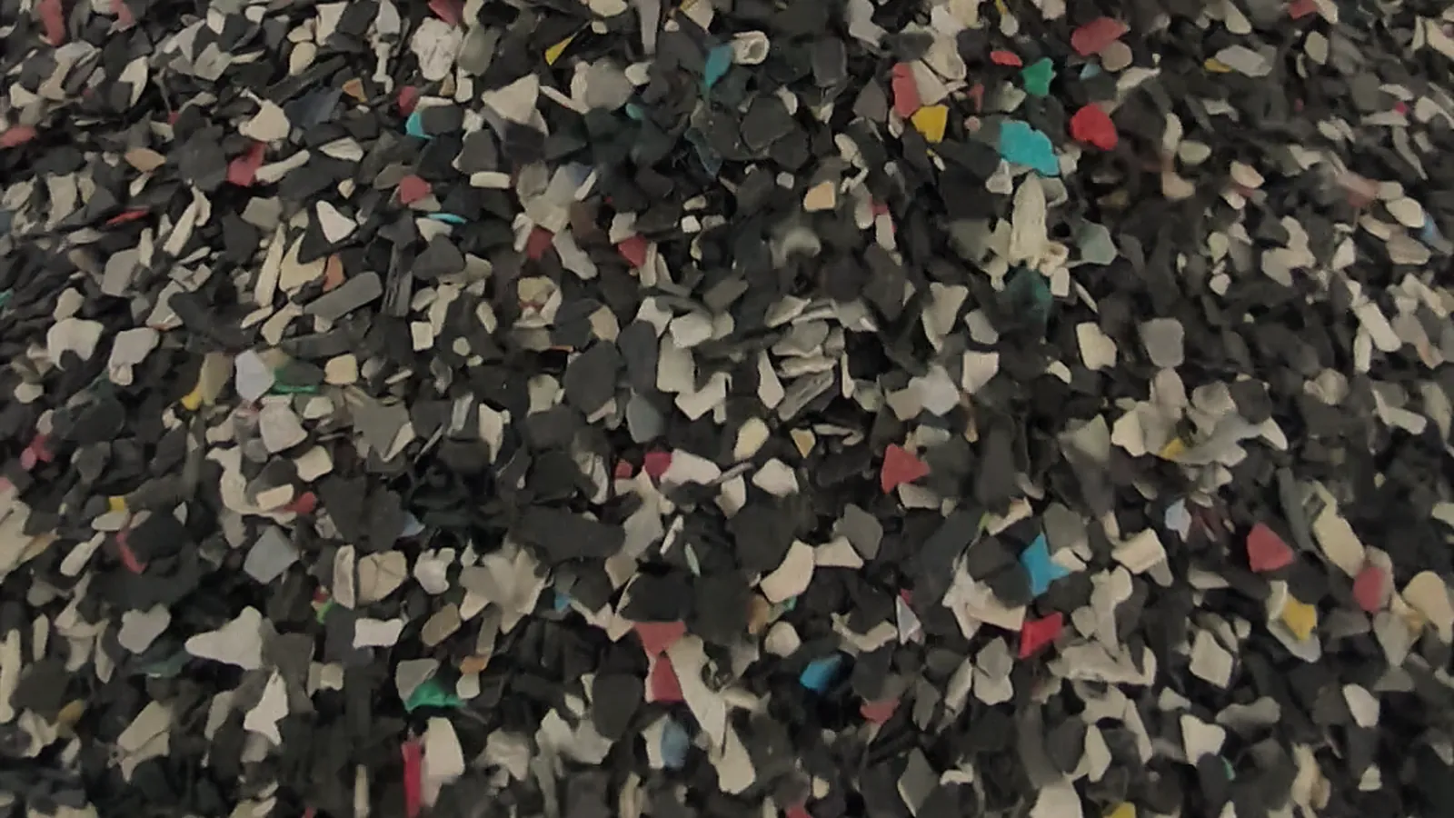 Crushed plastic flakes in mixed colours, mostly dark grey and off-white with flecks of red and teal, in Dayatas Profab's production footage
